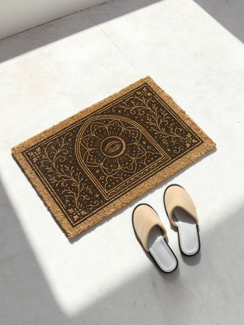 A rectangular, natural brown coir doormat with a non-slip black vinyl backing, viewed from directly above. The mat is made of textured coconut fiber. The design features intricate black line art in a Victorian Gothic and Art Nouveau style. In the center is an All Seeing Eye, or Eye of Providence, enclosed within a radiant starburst and a mandala-like floral pattern. This central motif is set inside an arched frame. The corners and sides are adorned with symmetrical, flowing botanical vines.
